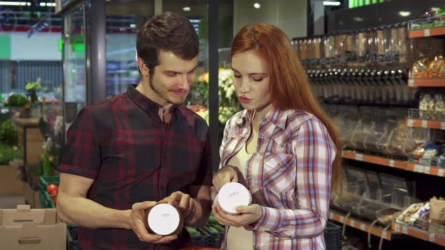 Attractive young couple arguing about the food composition data at the hypermarket. Handsome brunette man and his pretty redhead girlfriend pointing their forefingers on the can with some product