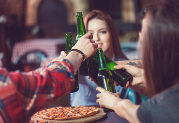 Friends having a drinks in a bar, They are sitting at a wooden table with beers and pizza. Focus on a gorgeous girl touching her bottle.