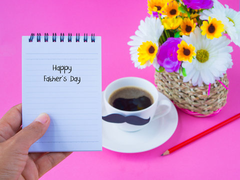 Father's day concept. Hand holding not book with Happy Father's Day message on note book with pink flower, coffee cup with black Mustache on pink background