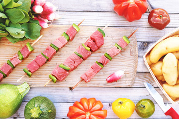 Portion of raw pork or beef on skewers, colorful tomatoes, radish, potato and young zucchini on wooden table ready for cooking. 