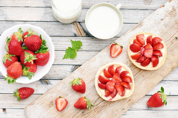 Homemade strawberry tarts on wooden board, cup of milk and bowl of fresh strawberry on wooden table. Countryside summer breakfast concept. 