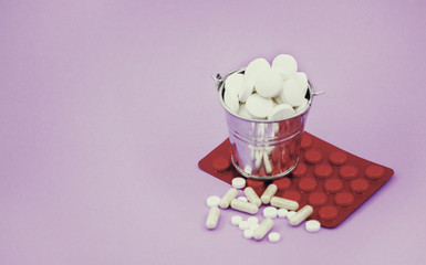 A full bucket of round tablets and a red packing of pills on a lilac background