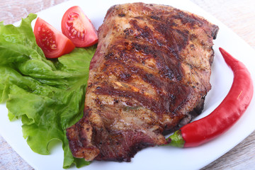 Pork ribs on grill and chili pepper with tomato, lettuce leaves on white plate