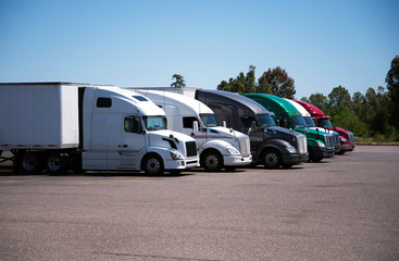 Semi-trucks and trailers of different make and models stand in row on flat parking lot of truck stop
