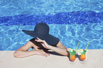 Beautiful woman in bikini and large hat relaxing at pool with cocktails. Summer holidays, travel, vacation concept. Copy space.