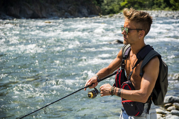 Young handsome male tourist standing and fishing in river in sunny day with rod