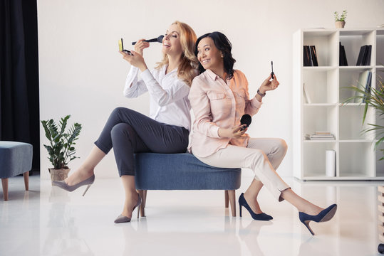 Two Smiling Middle Aged Businesswomen Sitting Together And Applying Makeup In Office