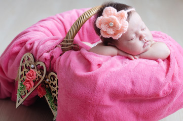 Newborn sleeping with knitted flower on head. Infant baby girl closeup lying on pink blanket in basket decorated with wooden hearts. Cute portrait of new child.