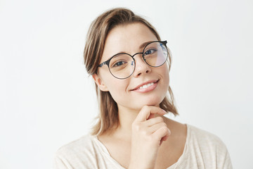 Portrait of young beautiful girl in glasses smiling looking at camera holding hand on chin over white background.