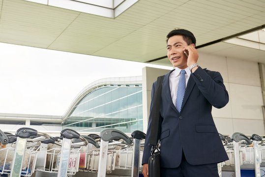 Portrait Of Modern Asian Businessman Speaking By Phone Leaving Airport With Luggage And Smiling