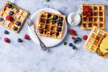 Homemade square belgian waffles with fresh ripe berries blueberry, raspberry, red currant, peach served with caramel, balsamic sauce, honey, whipped cream over gray background. Top view with space