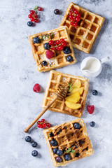 Homemade square belgian waffles with fresh ripe berries blueberry, raspberry, red currant, peach served with caramel, balsamic sauce, honey, whipped cream over gray background. Top view with space - obrazy, fototapety, plakaty