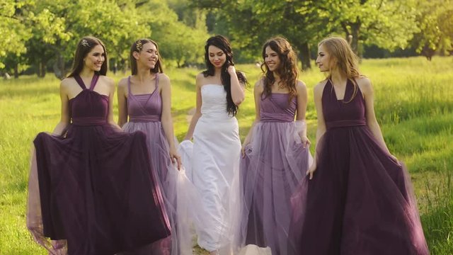 Beautiful bride and bridemaids in purple dresses walking in the park or garden holding hands and laughing in the evening.