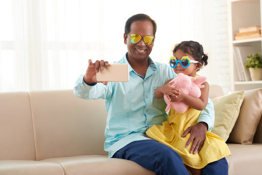 Smiling father and his cute little daughter wearing sunglasses taking selfie on smartphone while having fun in cozy living room