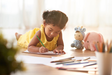 Concentrated toddler in yellow dress coloring picture with pencils while lying on floor, blurred background - obrazy, fototapety, plakaty