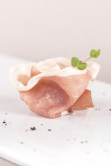 Fresh appetizer with prosciutto ham and slices of parmesan, decorated with green leaf, placed on white plate, light background, isolated