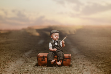 Little boy sitting on an old luggage in a vintage suit and holding wheat, against blurry background