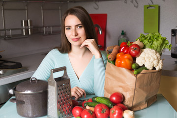 Young positive smiling  woman housewife in kitchen having many green vegetables in package on table, holding phone and searching recipe for cooking something in phone