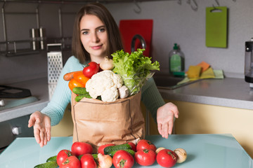 Young positive smiling woman housewife in kitchen having many green vegetables in package on table, holding phone and searching recipe for cooking something in phone