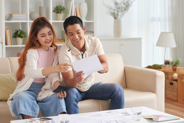 Obraz na płótnie Canvas Portrait of young Asian couple planning family budget in new house, smiling and talking sitting on sofa