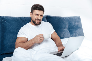 Young man working on laptop and having morning coffee in bed