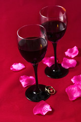Glasses with red wine, rose petals and wedding rings on a red sheet