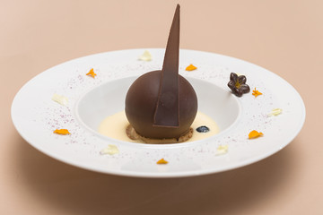 Luxurious dessert with chocolate globe and vanilla sauce, served on a biscuit, decorated with chocolate flower, flower petals and forest fruits, served in a white plate, light background, isolated