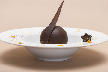 Luxurious dessert with chocolate globe and vanilla sauce, served on a biscuit, decorated with chocolate flower, flower petals and forest fruits, served in a white plate, light background, isolated