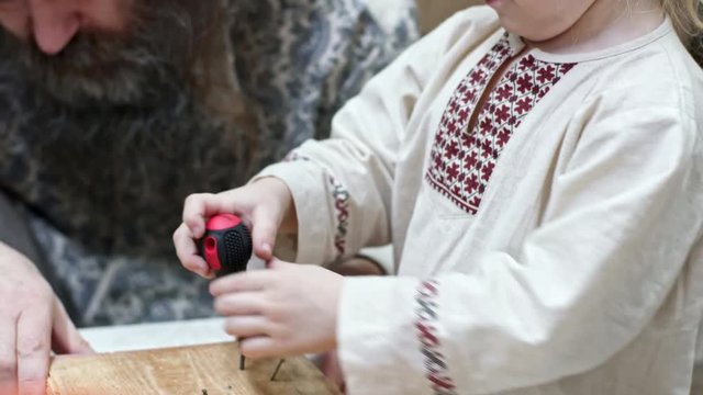 Blonde little boy in traditional Russian clothing driving nail into wood with screw driver with assistance of father