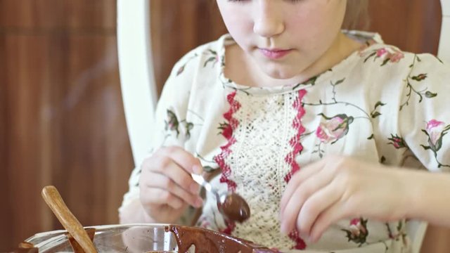 Tilt down of beautiful little child in traditional Russian dress pouring melted chocolate in silicone molds while cooking at kitchen table