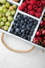 Assorted summer fruits and berries in a light wooden box with cells standing on light concrete background. Blueberries, raspberries, gooseberries, cherries. Top view