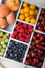 Assorted summer fruits and berries in a light wooden box with cells standing on light concrete background. Blueberries, raspberries, gooseberries, peaches, cherries. Top view