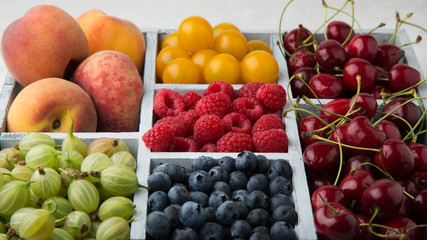 Naklejka na ściany i meble Assorted summer fruits and berries in a light wooden box with cells standing on light concrete background. Blueberries, raspberries, gooseberries, peaches, cherries, alycha