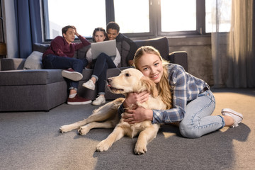 Smiling teenage girl having fun with golden retriever dog while friends sitting on sofa