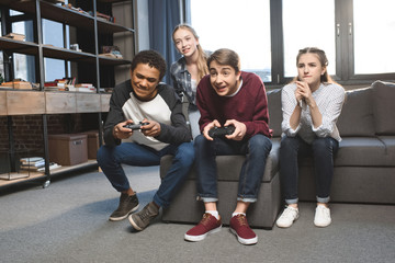 Fototapeta premium szczęśliwych wielokulturowych nastolatków grających w gry wideo z joystickami w domu, nastolatki bawiące się koncepcją