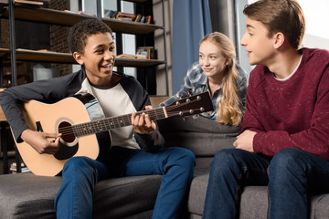 african american boy playing acustic guitar while his friends listening at home, teenagers playing...
