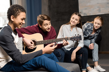 boy playing acustic guitar and singing while his friends listening at home, teenagers playing guitar concept