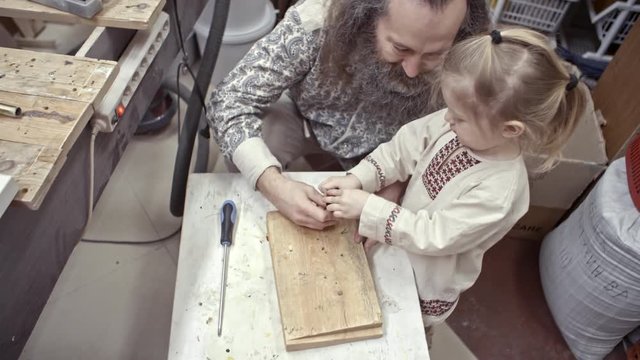 Top view of senior bearded man in traditional Russian clothing and his little son using screwdriver together while working in carpentry workshop