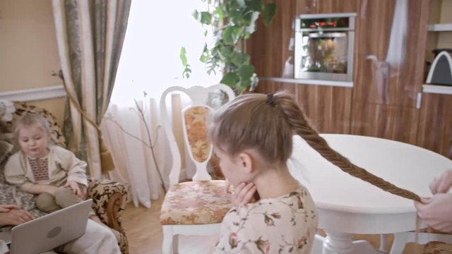 PAN of mother doing fishtail braid on hair of little daughter; boy and girl watching something on laptop with dad. Family wearing traditional Russian clothing at home