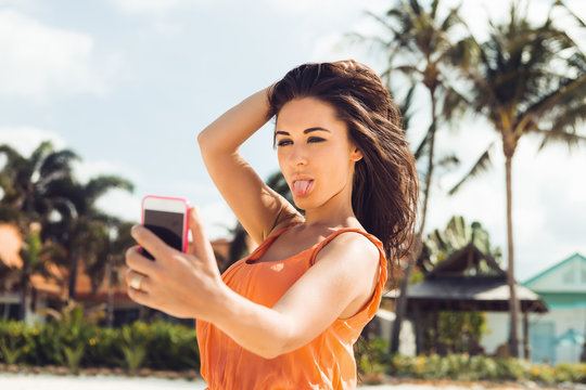 Close up of sexy hipster lady in light dress, sunglasses posing for selfie self portrait on a smartphone digital photocamera on a tropical beach. Swag urban teen girl. Outdoor lifestyle portrait.