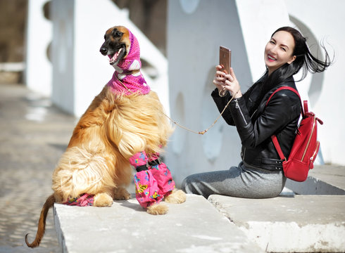 A girl in a city Park in the leather jacket with the red backpack laughs and takes photos with her phone of the Afghan hound. woman and dog walk