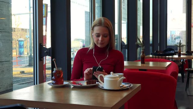 A young attractive woman sits at a table with meal in a cafe and listens to music on a smartphone while looking out a window with a smile