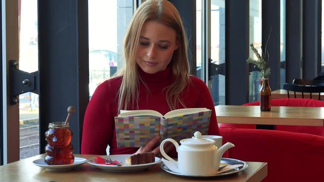 A young attractive woman sits at a table with meal in a cafe and reads a book with a smile