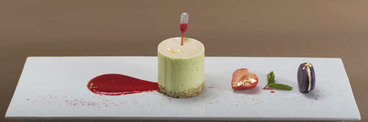 Luxurious dessert with pistachio mousse placed on biscuit, strawberry sauce served in a pipette, strawberry, macarons, mint, decorated with gold leafs – on a white plate, brown background