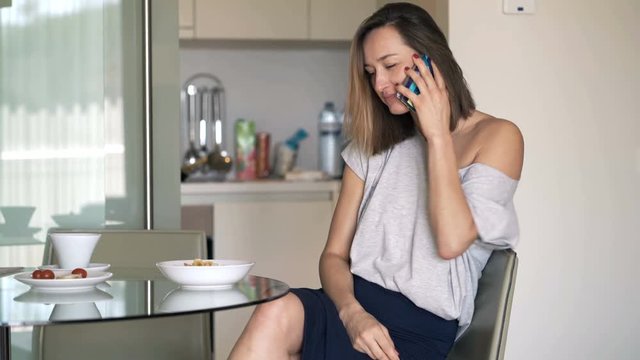 Young woman talking on cellphone and eating snack sitting in kitchen at home
