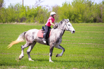 Young girl in a helmet riding a dapple-grey horse on a grass field