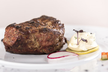 Succulent beef tenderloin steak, served with butter with herbs, decorated with radish and red and green herbs, placed on a white plate, light background, isolated