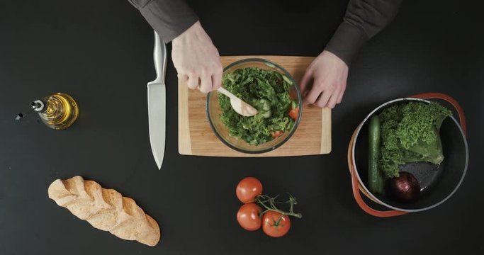 Man is making a salad in a bif transparent bowl. Other vegetables and bread placed nearby.
