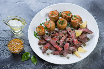 Grilled tagliata beef steak served with baked potatoes on a white plate, studio shot on a grey stone background