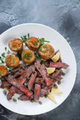 Top view of a white plate with tagliata beefsteak and baked potato on a grey stone surface, vertical shot